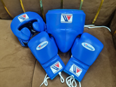 Official Winning professional 3-piece boxing sparring gear set in a matching classic blue premium leather colorway, featuring lace-up training gloves, a face guard headgear, and a groin protector grouped together on a brown surface.