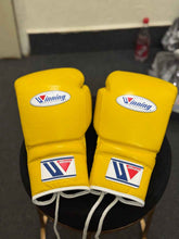 Winning professional lace-up boxing gloves in vibrant yellow leather with official white branding patches and white laces resting on a stool.