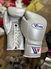 Custom Winning professional lace-up boxing gloves in metallic silver leather with official white branding patches and white laces resting on a stool.