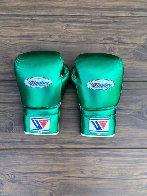 Custom Winning professional 3-piece boxing sparring gear set in a matching shimmering metallic green premium leather colorway, featuring lace-up training gloves, a face guard headgear, and a groin protector arranged on a wooden floor.