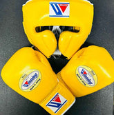 Official Winning professional boxing sparring set in a matching classic yellow premium leather colorway, featuring lace-up training gloves and a face guard headgear over a dark backdrop.