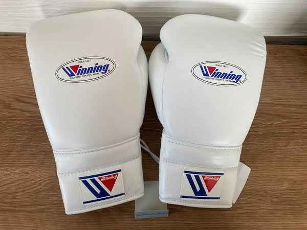 Official Winning professional lace-up training boxing gloves in pristine white premium leather resting on a wooden surface, featuring the iconic brand logo oval crest on the knuckle face and cuff patches.