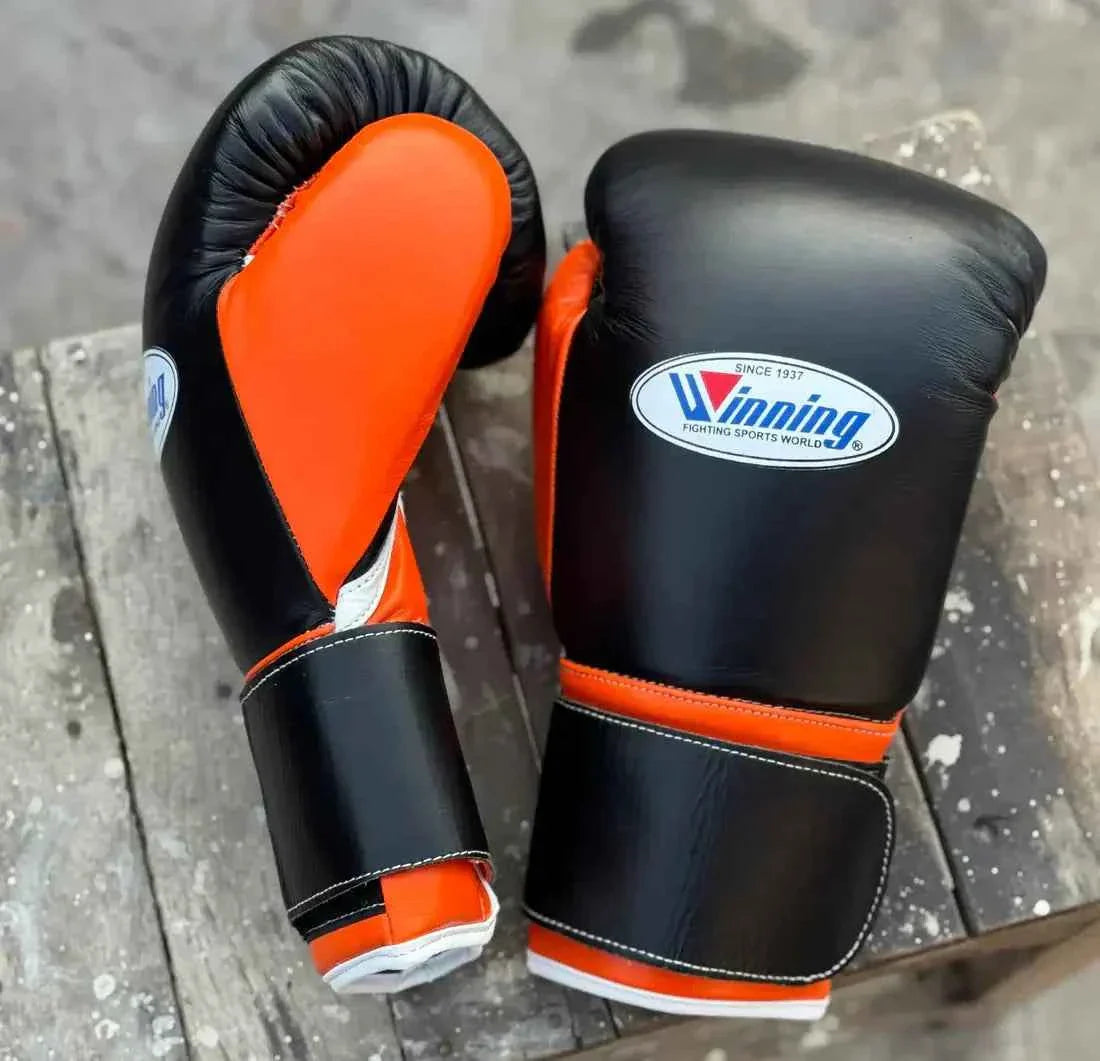 Custom Winning professional hook-and-loop boxing gloves in black leather featuring bright orange thumb linings, orange wrist borders, official white brand patches, and wide velcro straps.