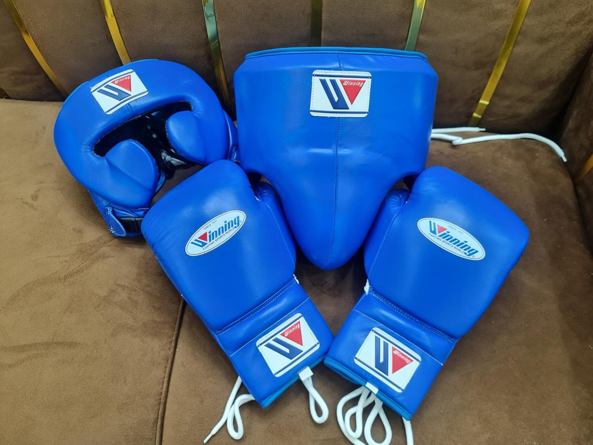 Official Winning professional 3-piece boxing sparring gear set in a matching classic blue premium leather colorway, featuring lace-up training gloves, a face guard headgear, and a groin protector grouped together on a brown surface.
