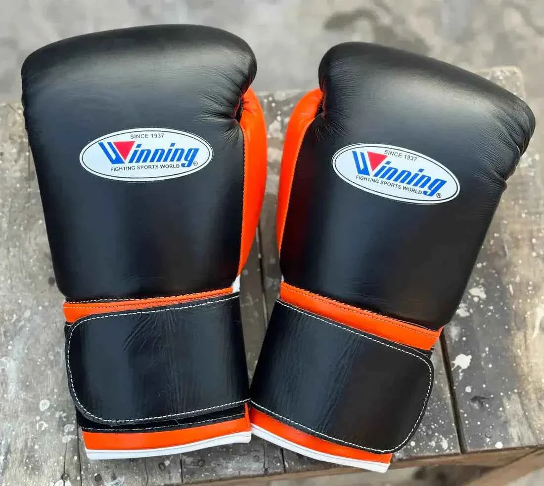 Custom Winning professional hook-and-loop boxing gloves in black leather featuring bright orange thumb linings, orange wrist borders, official white brand patches, and wide velcro straps.