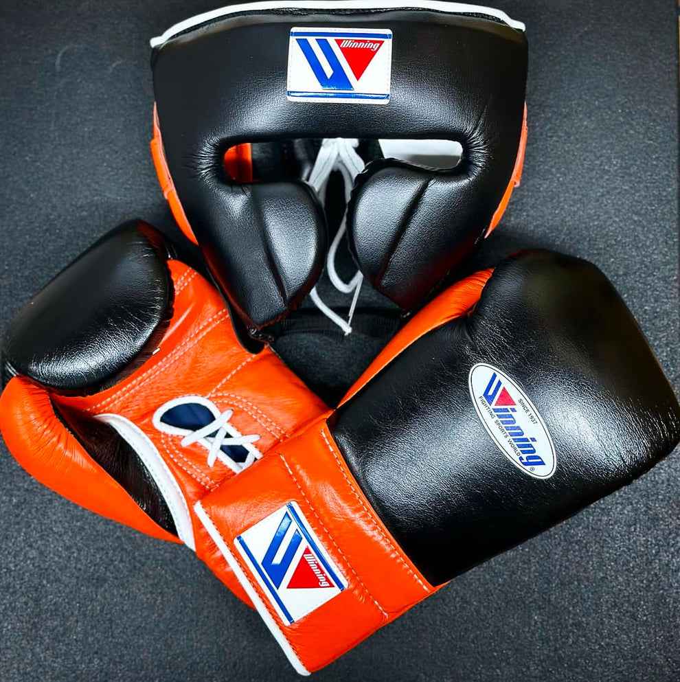 Custom Winning professional boxing sparring set in a matching black and orange premium leather colorway, featuring lace-up training gloves and a face guard headgear with white laces and brand crests.