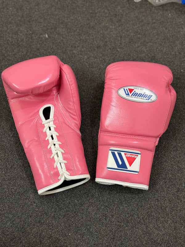 Official Winning professional lace-up training boxing gloves in premium pink leather resting on a dark stool, featuring white laces, white contrast edge trim, and the iconic brand logo crest on the cuffs.