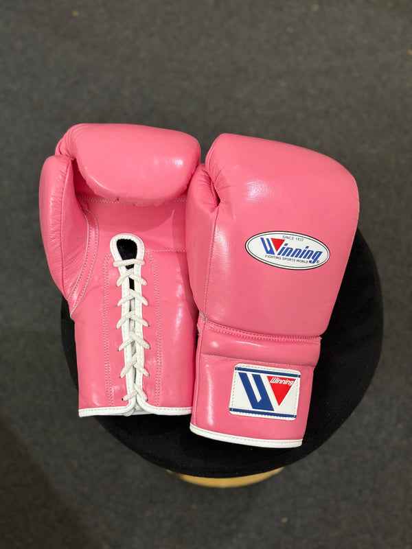Official Winning professional lace-up training boxing gloves in premium pink leather resting on a dark stool, featuring white laces, white contrast edge trim, and the iconic brand logo crest on the cuffs.