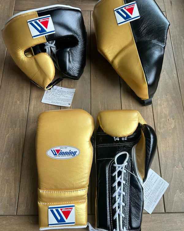 Custom Winning professional 3-piece boxing sparring gear set featuring metallic gold and black leather split-color designs across the lace-up training gloves, face guard headgear, and groin protector arranged on a wooden floor.