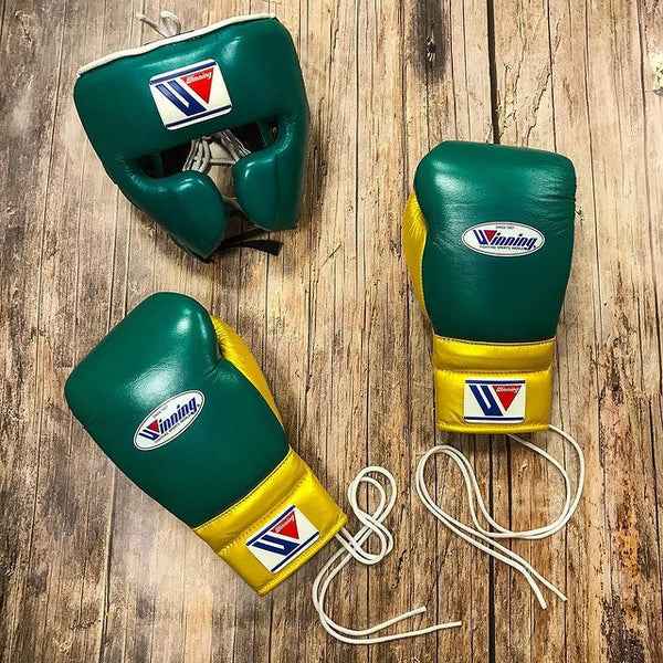 Custom Winning professional boxing sparring set in a matching forest green and yellow premium leather colorway, featuring lace-up training gloves and a face guard headgear resting on a rustic wooden floor.